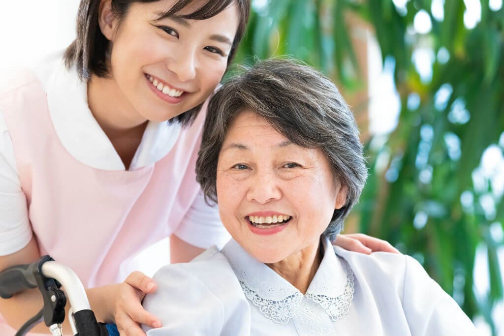 笑顔の車いすの女性とその肩に手を置く介護士の女性の写真